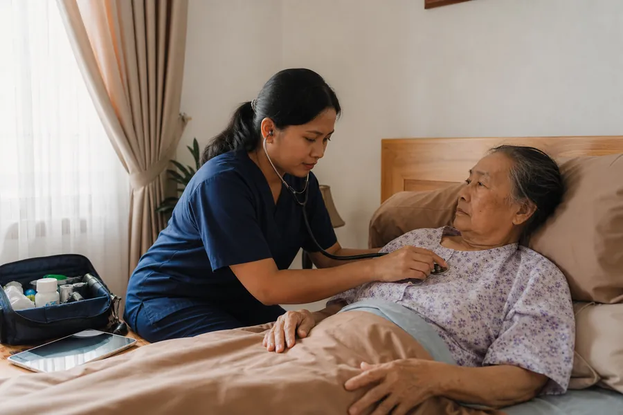 Perawat memeriksa seorang ibu lansia di tempat tidur dengan stetoskop